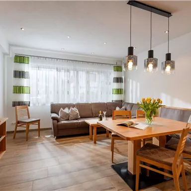 Ein gemütlicher Wohnraum mit einem großen Tisch, Stühlen und einer komfortablen Couch. Die Fenster lassen viel Tageslicht herein und die Dekoration ist modern und stilvoll.