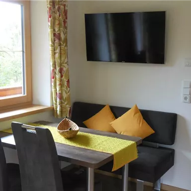 A cozy dining area with a table, two chairs, and a bench. A TV hangs on the wall and the windows are decorated with colorful curtains.