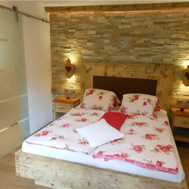 A cozy bedroom with a double bed and floral bedding. The walls are decorated with stone and there are warm light sources.