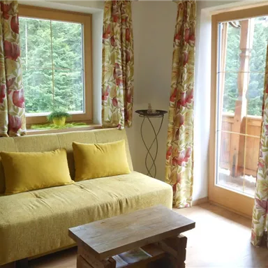 A bright living room with a yellow sofa and a wooden coffee table. Large windows provide a view of nature.