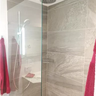 A modern shower with a glass cabin and gray tiles. Red towels hang on the wall, adding a colorful accent to the room.