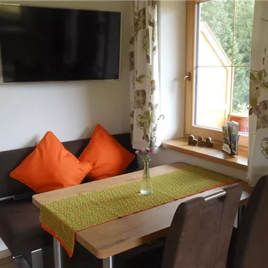 A cozy dining area with a wooden table and chairs. The wall is equipped with a television, and there is a large window with curtains.