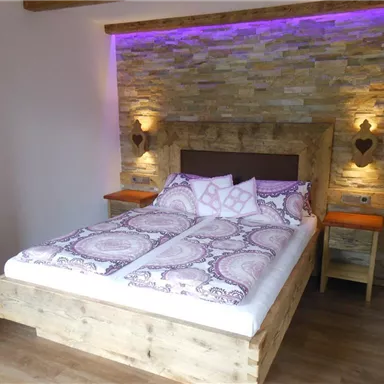 A cozy bedroom with a large bed and a rustic wooden frame. The walls are made of stone and soft purple light creates a relaxed atmosphere.