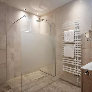 A modern bathroom with a walk-in shower and elegant tiles.
Towels hang on a towel holder and a sink with a wooden base cabinet is visible.