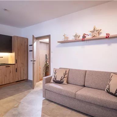 A modern living room with a beige sofa and a wooden wall shelf. The kitchen in the background is made of wood and well-lit.