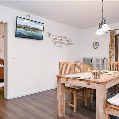A bright dining room with a large wooden table and chairs. A television is hanging on the wall, and there is a door to the outdoor area.