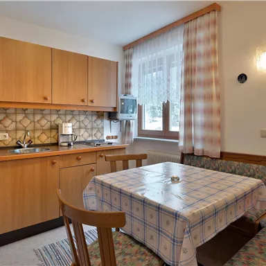 A cozy kitchenette with wooden cabinets and a dining area. The room is bright and cheerful with windows and soft cushions.