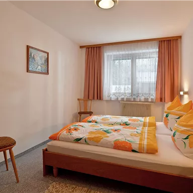 A cozy bedroom with two beds and colorful bedding. The room has a window with curtains and a chair in the corner.