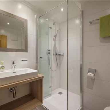 A modern bathroom with a shower and a sink. The towels are green, and the walls are light tiled.