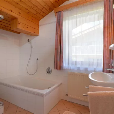 A bright bathroom with a window, a bathtub, and a shower. The wooden ceiling gives the room a cozy atmosphere.