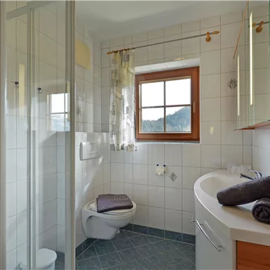 A modern bathroom with a shower and a window. It has bright tiles and wood details, as well as towels on a shelf.