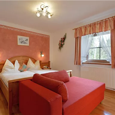 A cozy bedroom with a double bed and a red couch. The walls are painted in soft colors, with a window that lets in daylight.