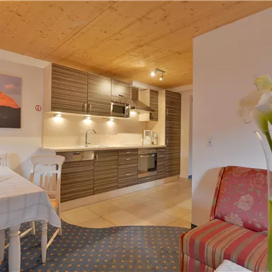 Eine moderne Küche mit Holzelementen und einem Essbereich. Im Vordergrund steht eine gemütliche Couch mit frischen Blumen.