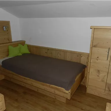 Ein einfaches Schlafzimmer mit einem Holzbett, grünem Kissen und einer Holzkommode. Der Raum hat einen gemütlichen und natürlichen Look.