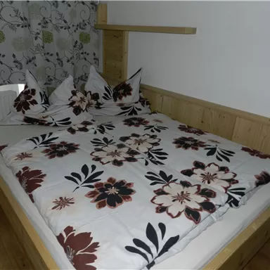 Ein gemütliches Bett mit floralem Bettzeug und Kissen. Im Hintergrund ist ein Vorhang mit dezentem Muster zu sehen.