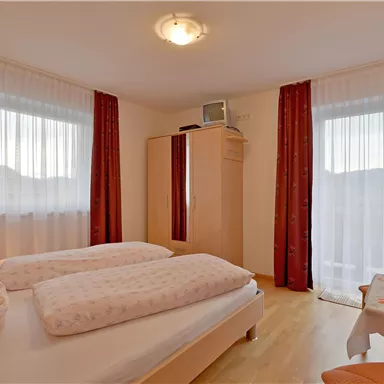 A bright bedroom with two single beds and large windows. The walls are in soft colors, and there is a wardrobe as well as a small table with a plant.