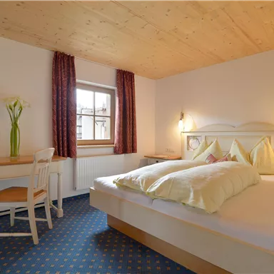 Ein gemütliches Schlafzimmer mit einem großen Bett und hellen Kissen. Der Raum hat einen Schreibtisch und eine Vase mit frischen Blumen.