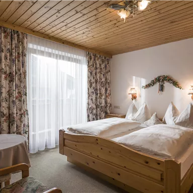 A cozy bedroom with wooden paneling and two single beds. Curtains with floral patterns and a small table complete the ambiance.