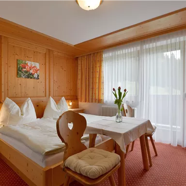 Ein gemütliches Schlafzimmer mit Holzvertäfelung und zwei Betten. Ein Tisch mit frischen Blumen steht am Fenster, das viel Tageslicht hereinlässt.