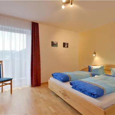 A cozy bedroom with two beds and blue bedspreads. A chair and a window with curtains create a bright, welcoming atmosphere.