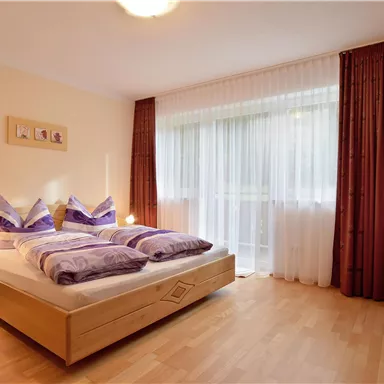 A cozy bedroom with a double bed and colorful pillows. The room has large windows with curtains and a television in the corner.
