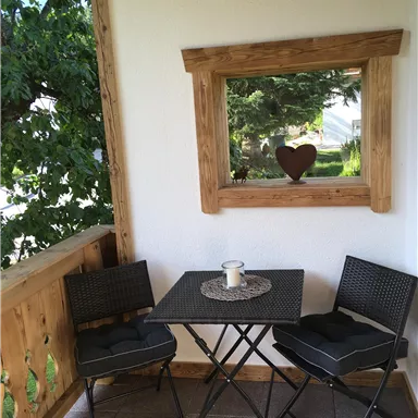 Ein kleiner Balkon mit zwei schwarzen Stühlen und einem Tisch. Im Hintergrund ist ein Fenster mit Blick ins Grüne und einem herzförmigen Dekor.