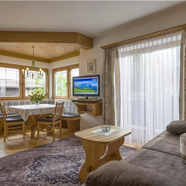 A cozy living room with a sofa and a wooden table. Large windows provide plenty of daylight and a view of nature.