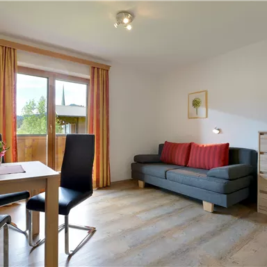 Ein gemütliches Wohnzimmer mit einem grauen Sofa, einem Esstisch und Stühlen. Große Fenster lassen viel Licht herein und bieten einen Blick nach draußen.