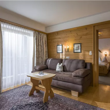 A cozy living ambiance with a brown sofa and a small table. The room has large windows with curtains and access to another room.