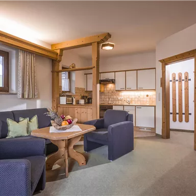 A cozy living room with blue armchairs and a wooden table. The open kitchen is modern and warmly furnished.