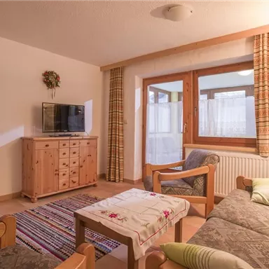 Ein gemütliches Wohnzimmer mit einer Couch, einem Fernseher und einem Holztisch. Große Fenster sorgen für viel Tageslicht und einen Blick auf den angrenzenden Raum.