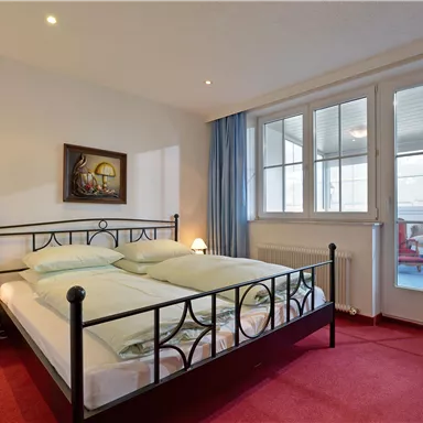 Ein helles Schlafzimmer mit einem Doppelbett und einem roten Teppich. Große Fenster sorgen für viel Tageslicht und eine angenehme Atmosphäre.