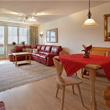Ein gemütliches Wohnzimmer mit einer roten Couch und einem Holztisch. Der Raum ist hell und einladend, mit großen Fenstern und dekorativen Vorhängen.