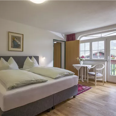 Ein gemütliches Schlafzimmer mit einem Doppelbett, zwei Kissen und einem Tisch mit Stühlen. Große Fenster sorgen für viel Tageslicht und bieten einen Blick nach draußen.