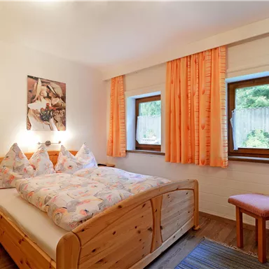 A cozy bedroom with a double bed and colorful pillows. The windows are decorated with orange curtains and there is a small sitting area.