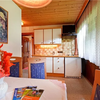 A cozy kitchen with a dining table, chairs, and a vase of flowers. Bright furniture and a window with curtains create an inviting atmosphere.