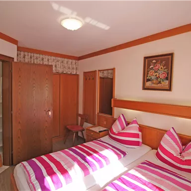 A cozy bedroom with two beds and colorful cushions. The room is bright and friendly with wooden paneling and a bathroom in the background.