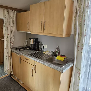 A small kitchenette with wooden cabinets and a countertop. It is equipped with a coffee machine and has windows with curtains.