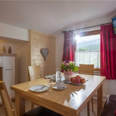 A cozy dining area with wooden chairs and a table on which fresh flowers and fruit are placed. Light streams through the window, creating an inviting atmosphere.