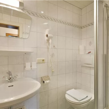 Ein modernes Badezimmer mit weißen Fliesen und einem Waschbecken. Es gibt eine Dusche, einen Spiegel und eine Toilette.