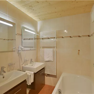 Ein modernes Badezimmer mit einer Badewanne und zwei Waschbecken. Es gibt helle Fliesen und eine Holzdecke, die eine gemütliche Atmosphäre schafft.