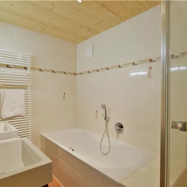 Ein modernes Badezimmer mit einer Badewanne und separater Dusche. Die Wände sind hell gefliest, und es gibt zwei Waschbecken.