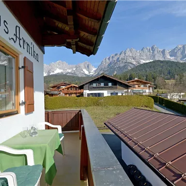 Ein Balkon mit einem Tisch und Stühlen, der eine schöne Aussicht auf die Berge bietet. Im Hintergrund sind weitere Häuser und eine klare, blaue Himmel zu sehen.