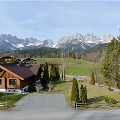 An idyllic landscape with a wooden house in the foreground and majestic mountains in the background. The scene features green fields and trees under a clear sky.