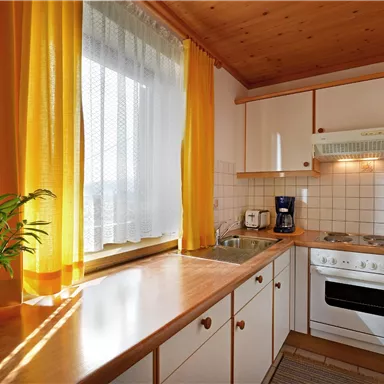 Eine helle Küche mit Holzoberflächen und orangefarbenen Vorhängen. Das Zimmer ist gut beleuchtet und hat eine Pflanzenarrangement auf der Arbeitsplatte.