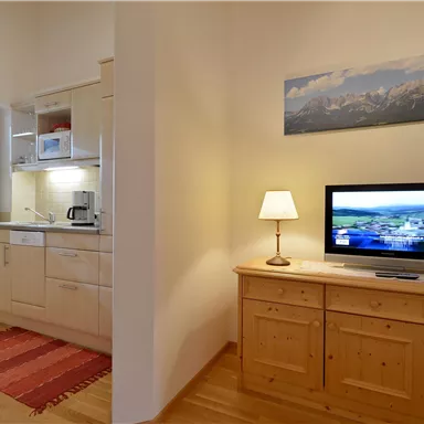 A modern kitchen with wooden cabinets and a small dining area. In the room, there is a television on a sideboard and a window that lets in natural light.