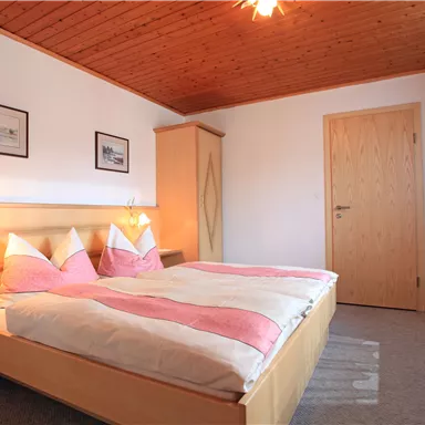 A cozy bedroom with a double bed and pink bedding. The walls are light, and there is a wooden ceiling with warm lighting.