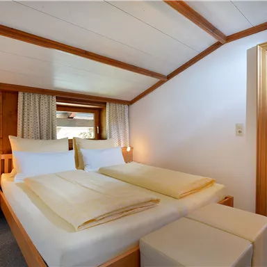 A cozy bedroom with two beds and warm wooden elements. There are windows with curtains and a pleasant lighting.