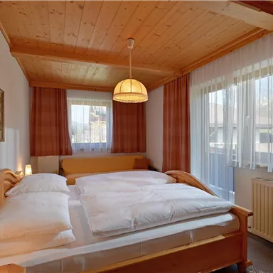 Ein gemütliches Schlafzimmer mit einem großen Doppelbett und Holzdecke. Helle Vorhänge lassen natürliches Licht herein und schaffen eine einladende Atmosphäre.