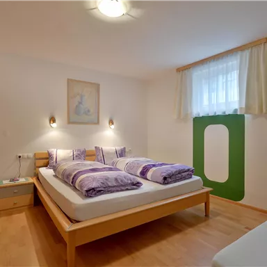 A cozy bedroom with two single beds and colorful cushions. The room has wooden flooring and large windows that provide plenty of daylight.